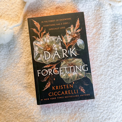 The book 'A Dark Forgetting' by Kristen Ciccarelli, featuring a dark cover with large white and gold floral illustrations, placed on a textured cream-colored blanket.