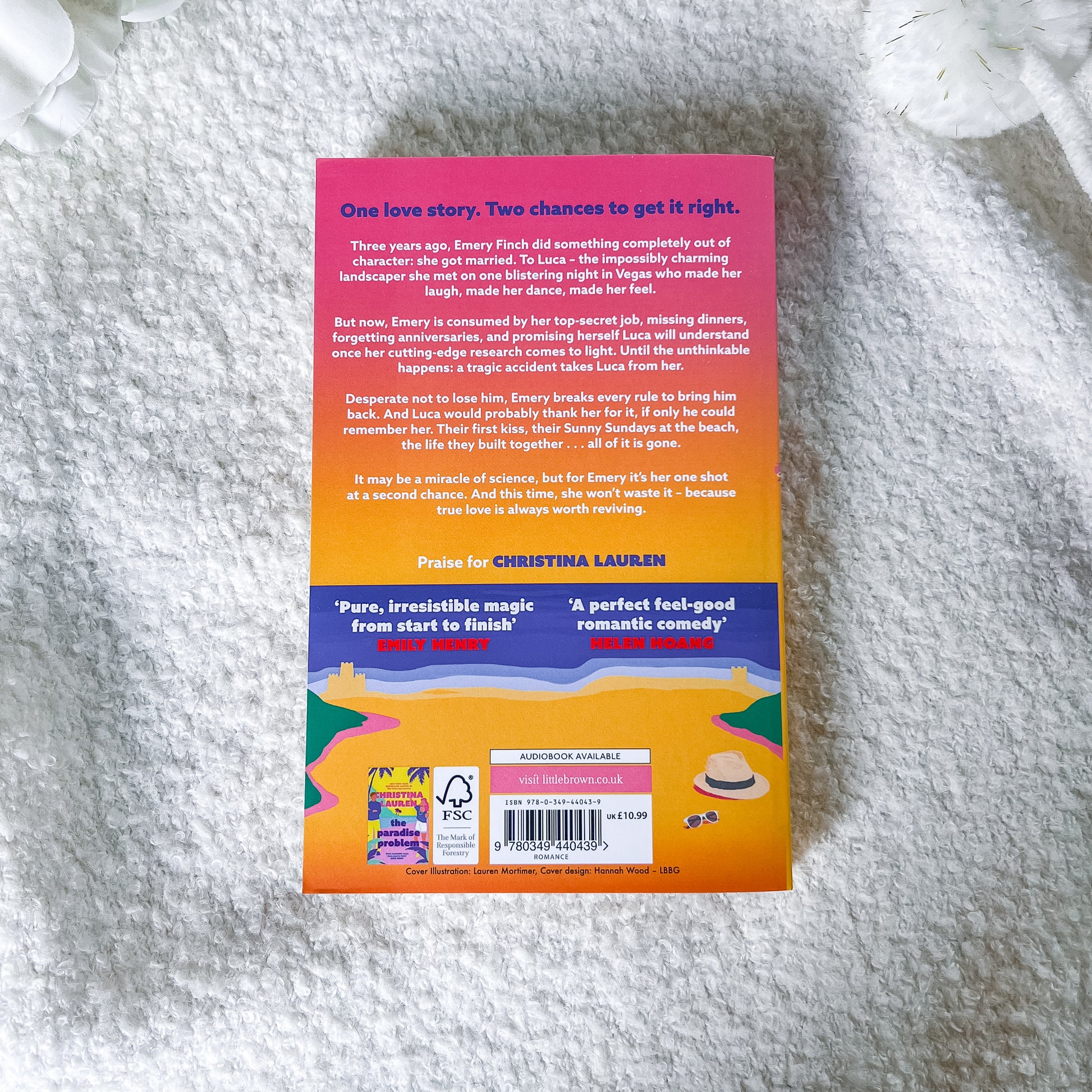 The back cover of the book by Christina Lauren is shown flat on a textured white fabric background. The cover features a vibrant gradient design transitioning from pink to purple and orange, with a cartoon illustration of a beach and a straw hat at the bottom.