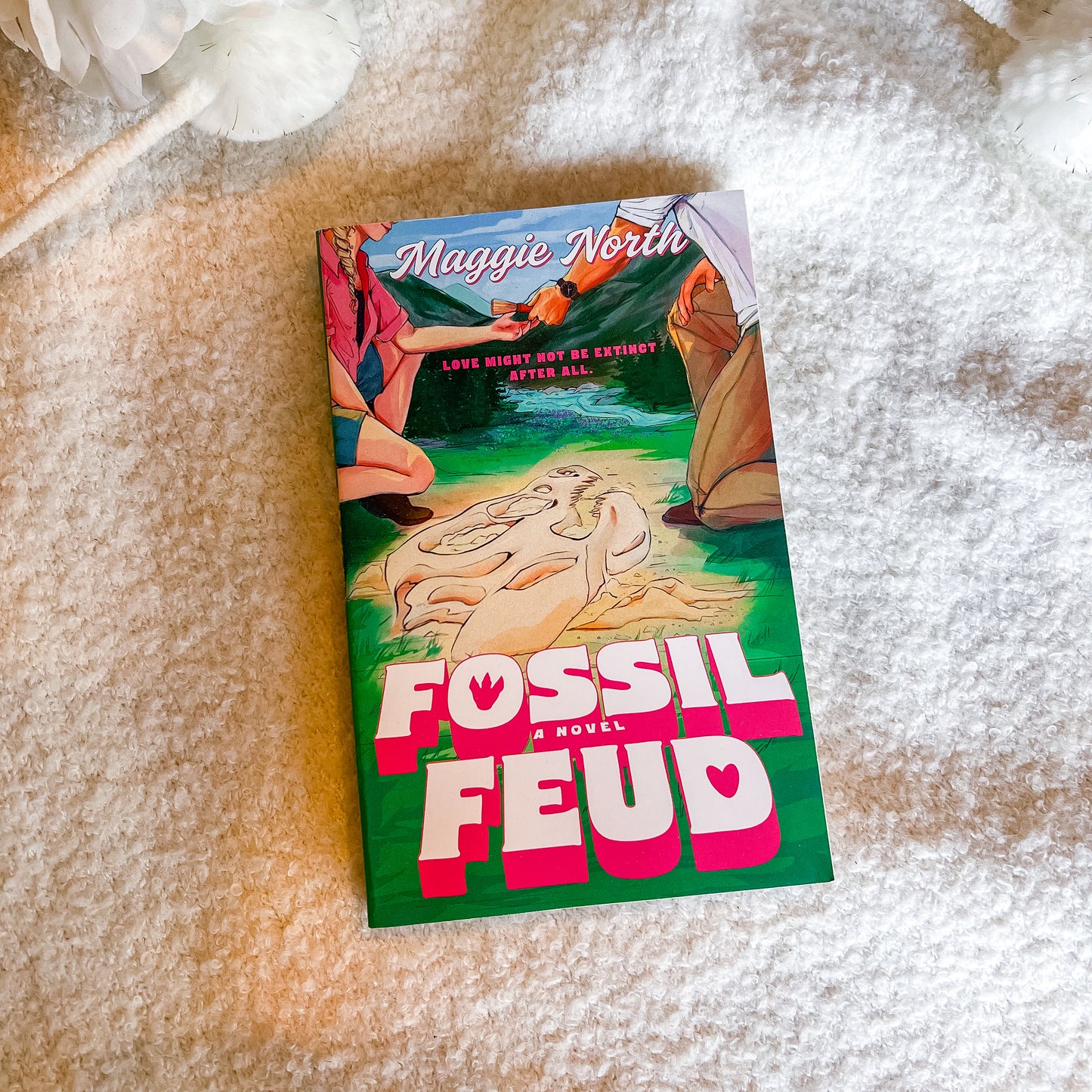 A paperback novel titled 'Fossil Feud' by Maggie North lies on a soft, textured cream-colored blanket. The cover features a colorful illustration with a green background, large pink and white title text, and a scene depicting a fossil discovery.