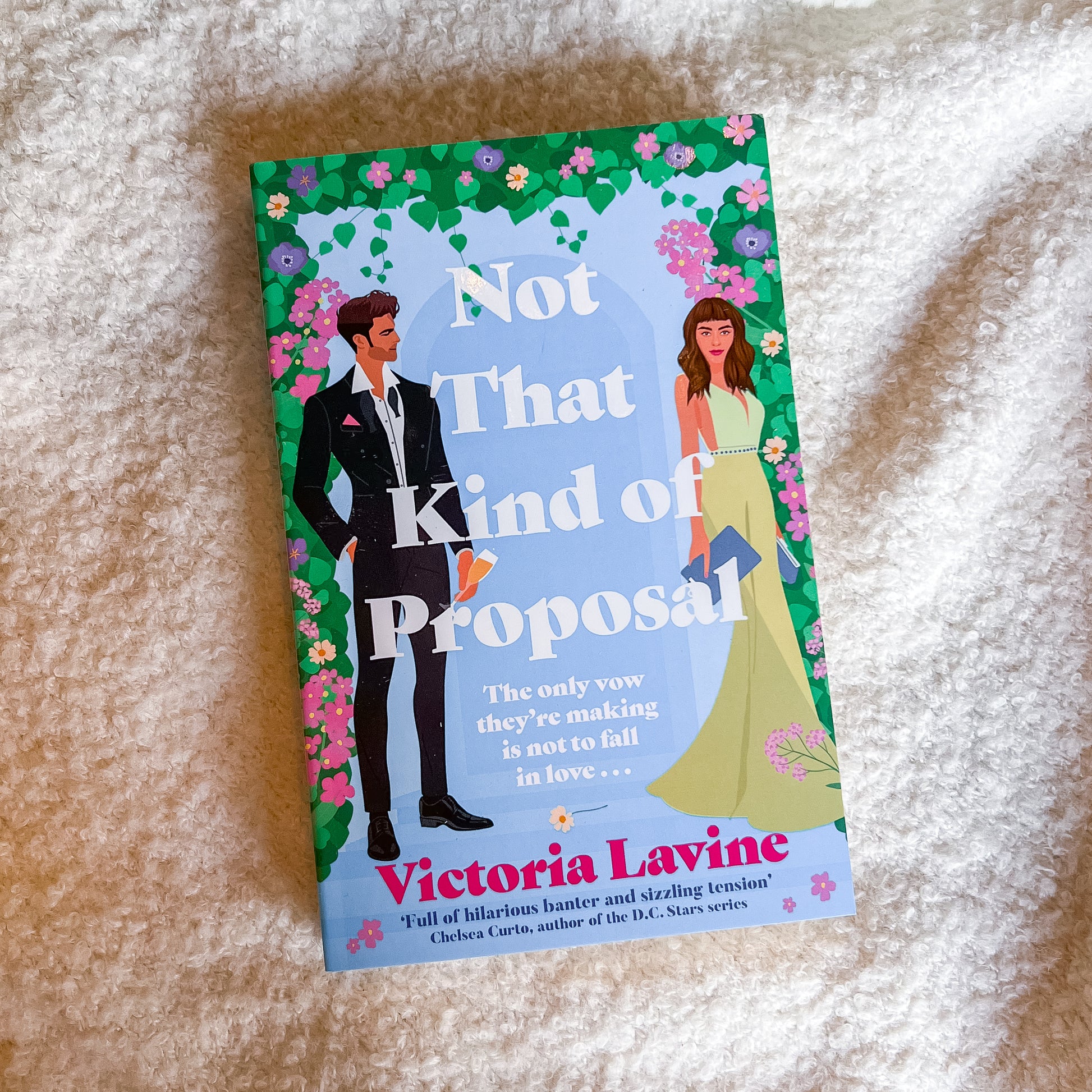 A paperback book titled 'Not That Kind of Proposal' by Victoria Lavine, featuring a colorful illustration of a man in a black suit and a woman in a yellow dress on a light blue background with a floral border, placed on a textured cream-colored blanket.