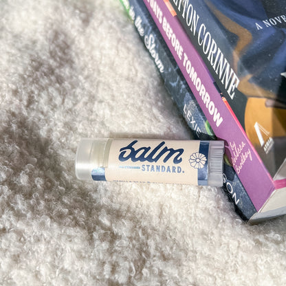 A tube of Balm Standard lip balm resting on a textured white faux fur surface next to a stack of books.