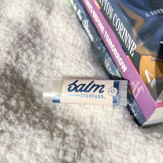 A tube of Balm Standard lip balm resting on a textured white faux fur surface next to a stack of books.