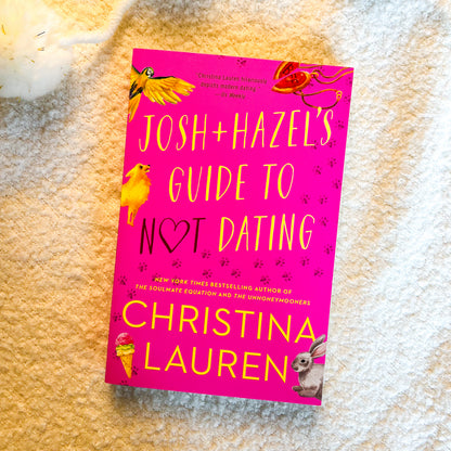 The book cover of 'Josh+Hazel's Guide to Not Dating' by Christina Lauren, featuring a bright pink background with yellow and black text and cartoon animal illustrations, photographed flat on a beige textured blanket.