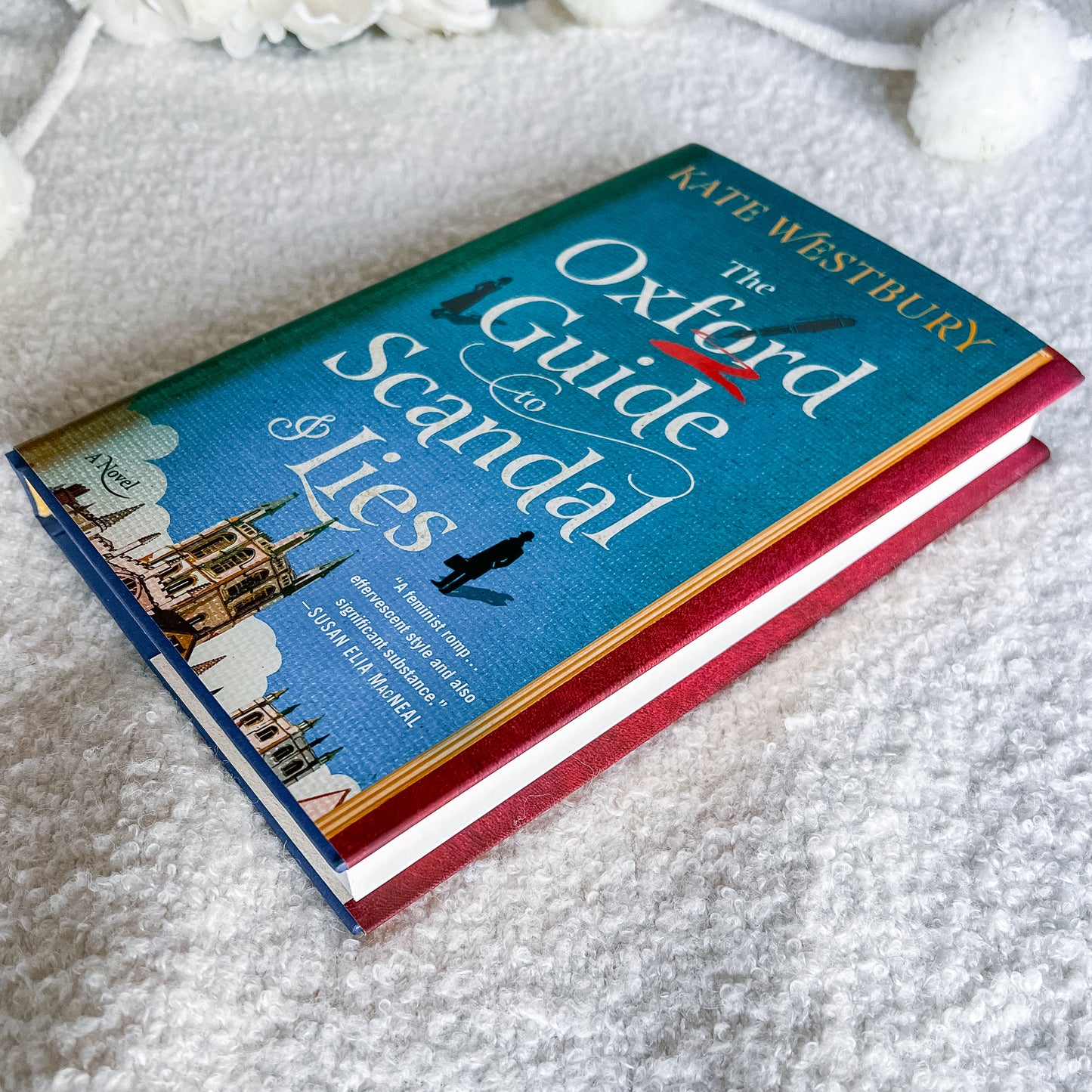 The book cover of 'The Oxford Guide to Scandal & Lies' by Kate Westbury, featuring a teal background with white title text, a red underline, and a small illustration of a cathedral at the bottom, shown on a pale pink background.