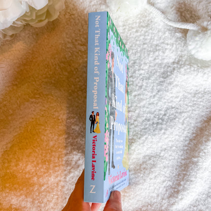 A hand holds a paperback book titled "Not That Kind of Proposal" by Victoria Lavine. The book cover features a pastel blue spine and front with pink floral illustrations and a small couple graphic.