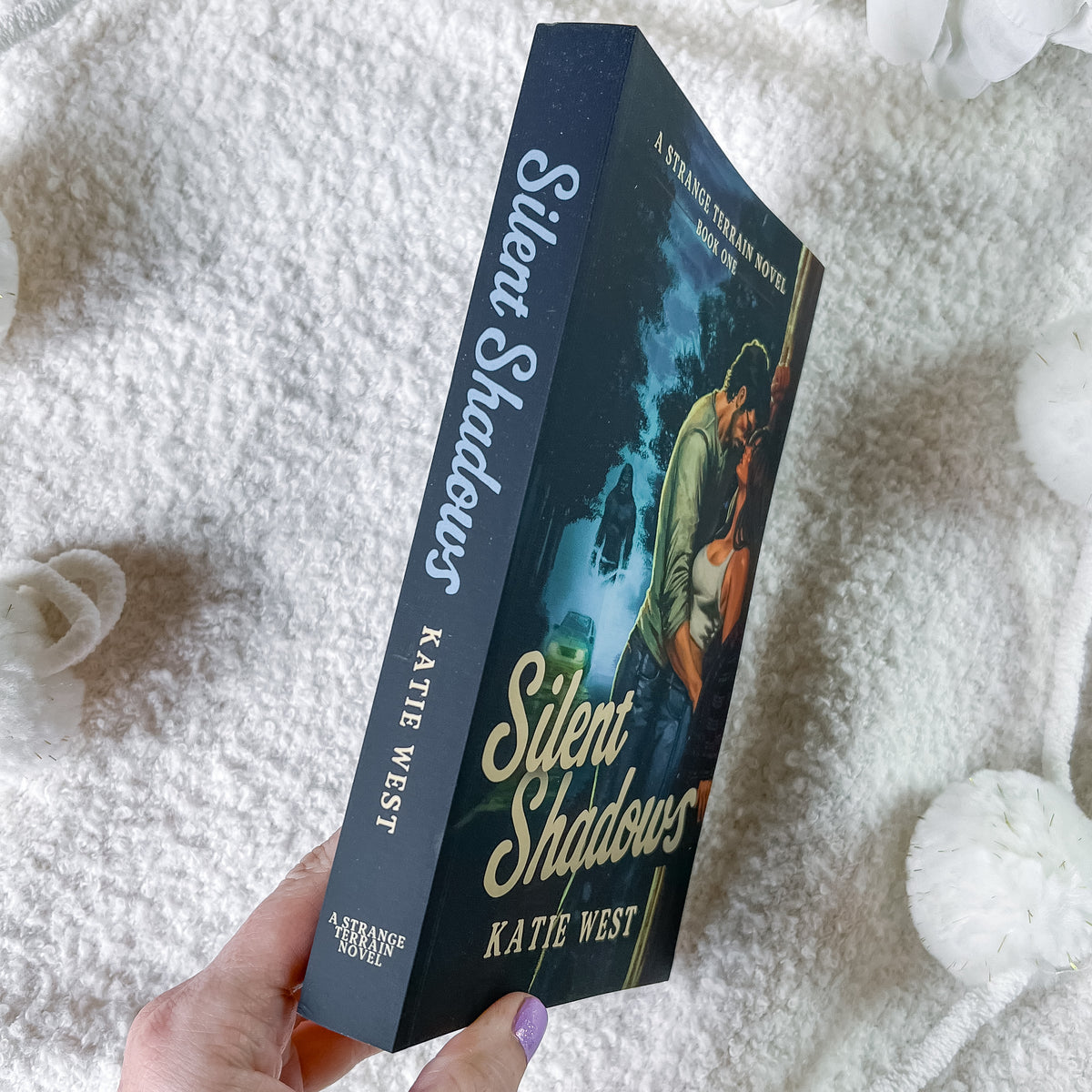 A hand holds a paperback copy of the novel 'Silent Shadows' by Katie West, set against a cozy white textured blanket background with decorative white pom-poms.