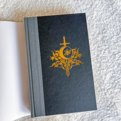 A hardcover with a dark charcoal gray cover featuring a gold foil emblem of a cross, crescent moon, and bees, shown open with blank white pages on a textured white background.