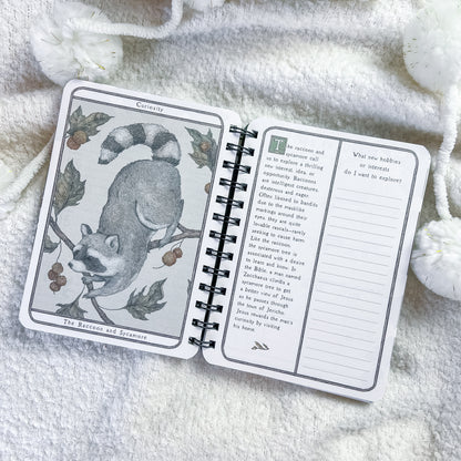 An open spiral-bound journal or notebook laid on soft white knit fabric. The left page shows an illustrated card titled 'Cats and Mice' with a raccoon on a branch, and the right page has text prompts for reflection and writing.