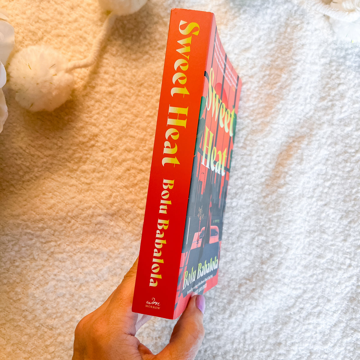 A hand holds a copy of the book 'Sweet Heat' , showing the bright red spine with yellow lettering, against a soft, textured cream blanket background.