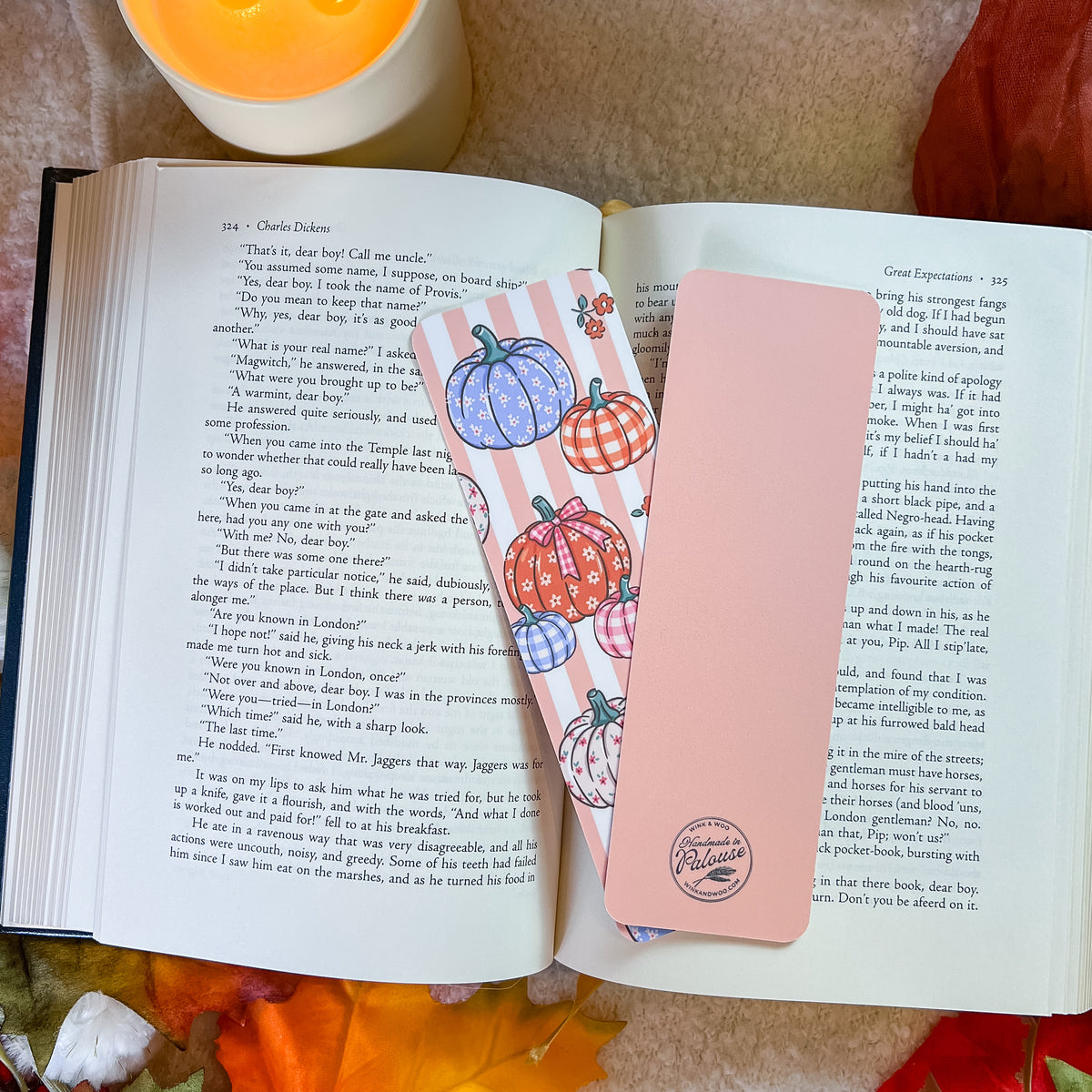 A pink bookmark with a pumpkin-themed design, styled on an open book with a warm blanket background.