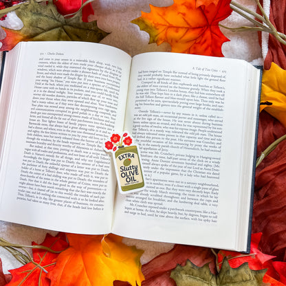 An open book spread photographed from above, surrounded by colorful autumn leaves. A small paper label on the right page reads 'EXTRA SLUTY OIL' with red floral graphics.