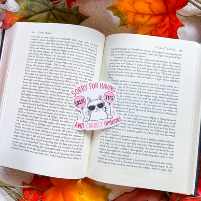 An open book spread with printed text, photographed from above on a surface with autumn leaves. A die-cut sticker on the pages features a cartoon cat wearing sunglasses and the text 'SORRY FOR HAVING GREAT TITS AND CORRECT OPINIONS'.