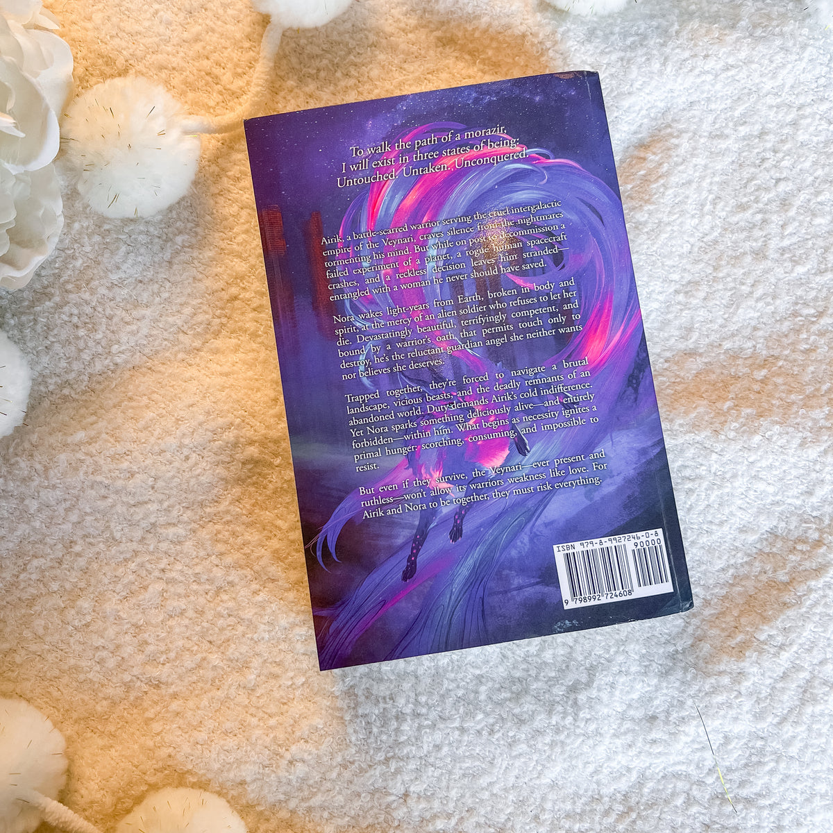 The back cover of a book, featuring a vibrant purple and pink illustration of a horse, laid flat on a textured white blanket with decorative white flowers.
