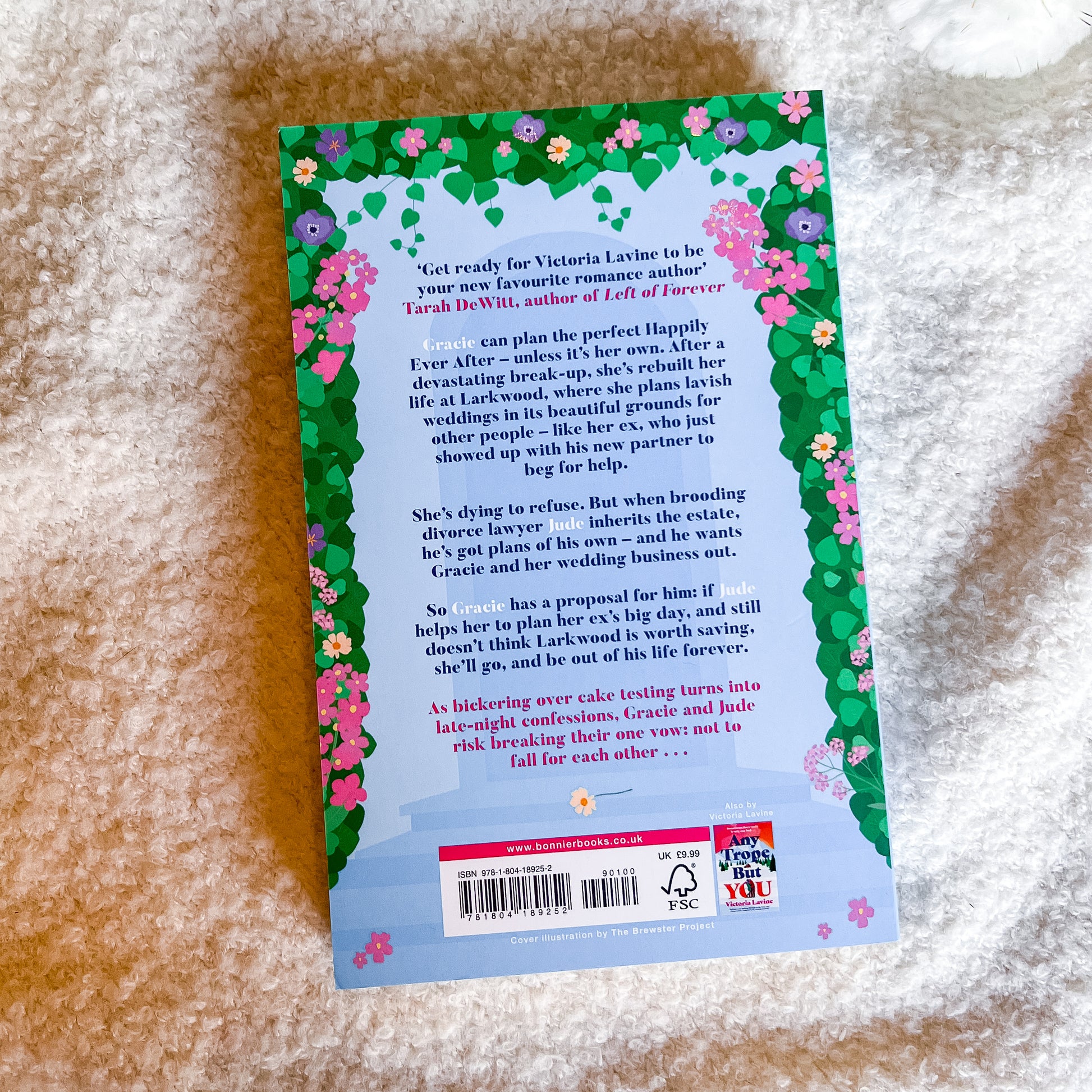 The back cover of a book titled 'Ever Ever After' by Sarah DeWitt, featuring a floral green border and a light blue illustration of a couple, photographed on a textured beige fabric background.