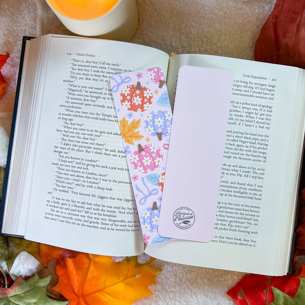A floral and autumn-themed bookmark rests on open book pages, surrounded by colorful fall leaves on a soft beige fabric background.