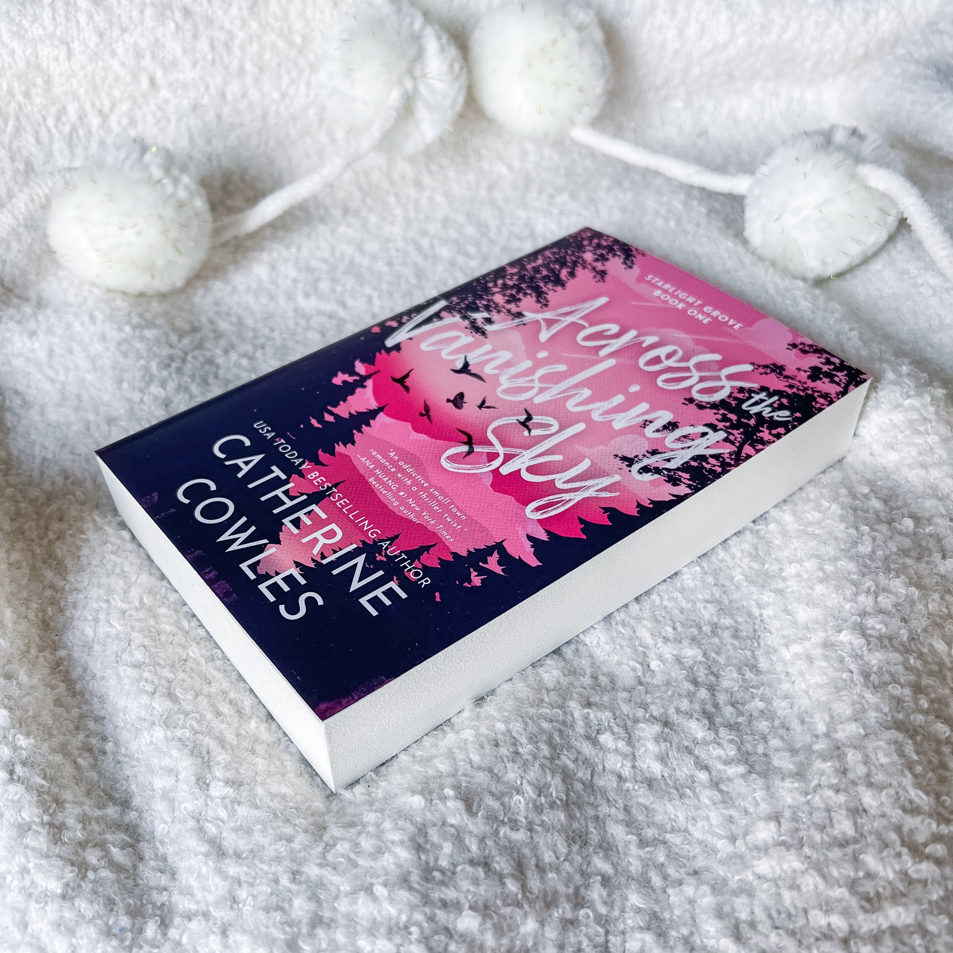 A book titled 'Across the Melting Sky' by Catherine Cowles lies on a soft white textured blanket with white pom-pom cords nearby. The cover is dark navy blue with pink floral artwork and white title text.