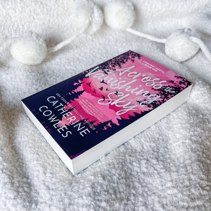 A book titled 'Across the Melting Sky' by Catherine Cowles lies on a soft white textured blanket with white pom-pom cords nearby. The cover is dark navy blue with pink floral artwork and white title text.