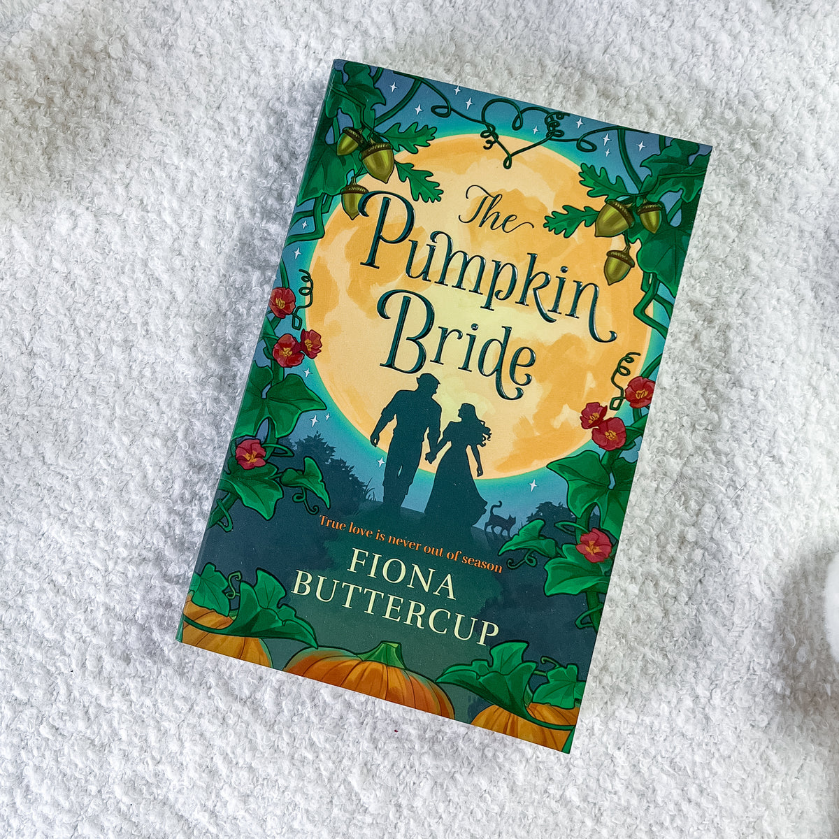 The book cover of 'The Pumpkin Bride' by Fiona Buttercup, featuring a festive autumn illustration with a large yellow moon, silhouetted couple, pumpkins, and red flowers, photographed on a white textured fabric background.