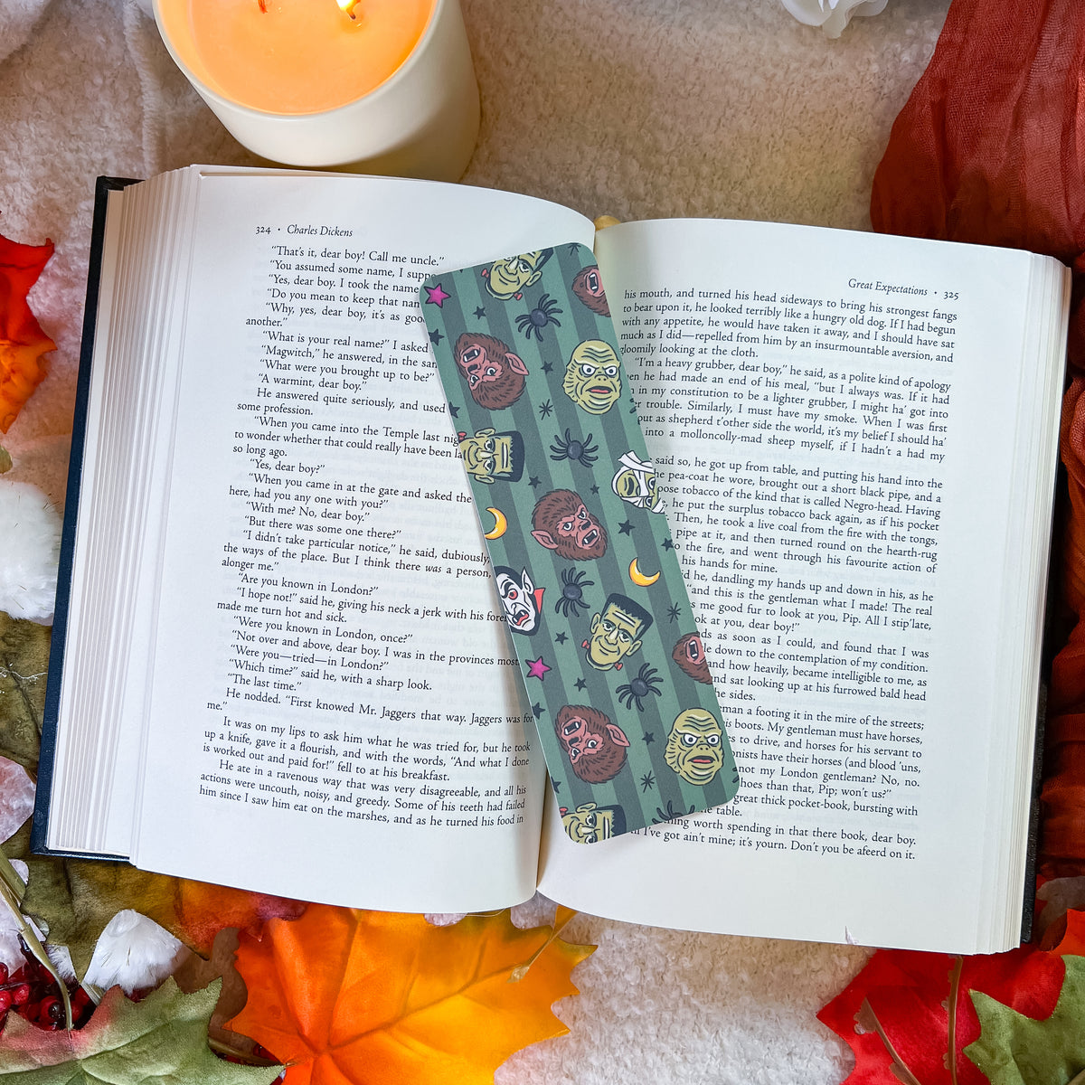 A decorative book bookmark with a teal background and colorful monster illustrations is shown inside an open book, styled with autumn leaves, a lit candle, and a cozy blanket.