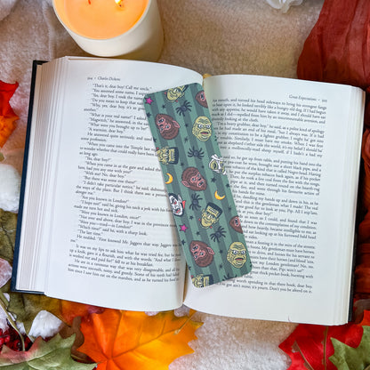 A decorative book bookmark with a teal background and colorful monster illustrations is shown inside an open book, styled with autumn leaves, a lit candle, and a cozy blanket.
