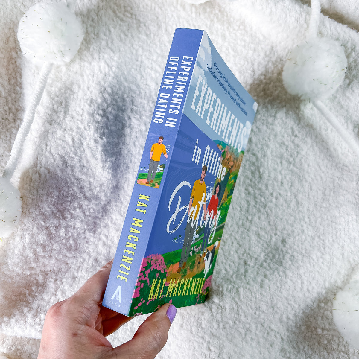 A hand holds a book titled 'Experiments in Offline Dating' by Kat MacKenzie. The book cover features a bright blue spine and front with colorful illustrations of people dating outdoors, set against a soft white textured blanket background.