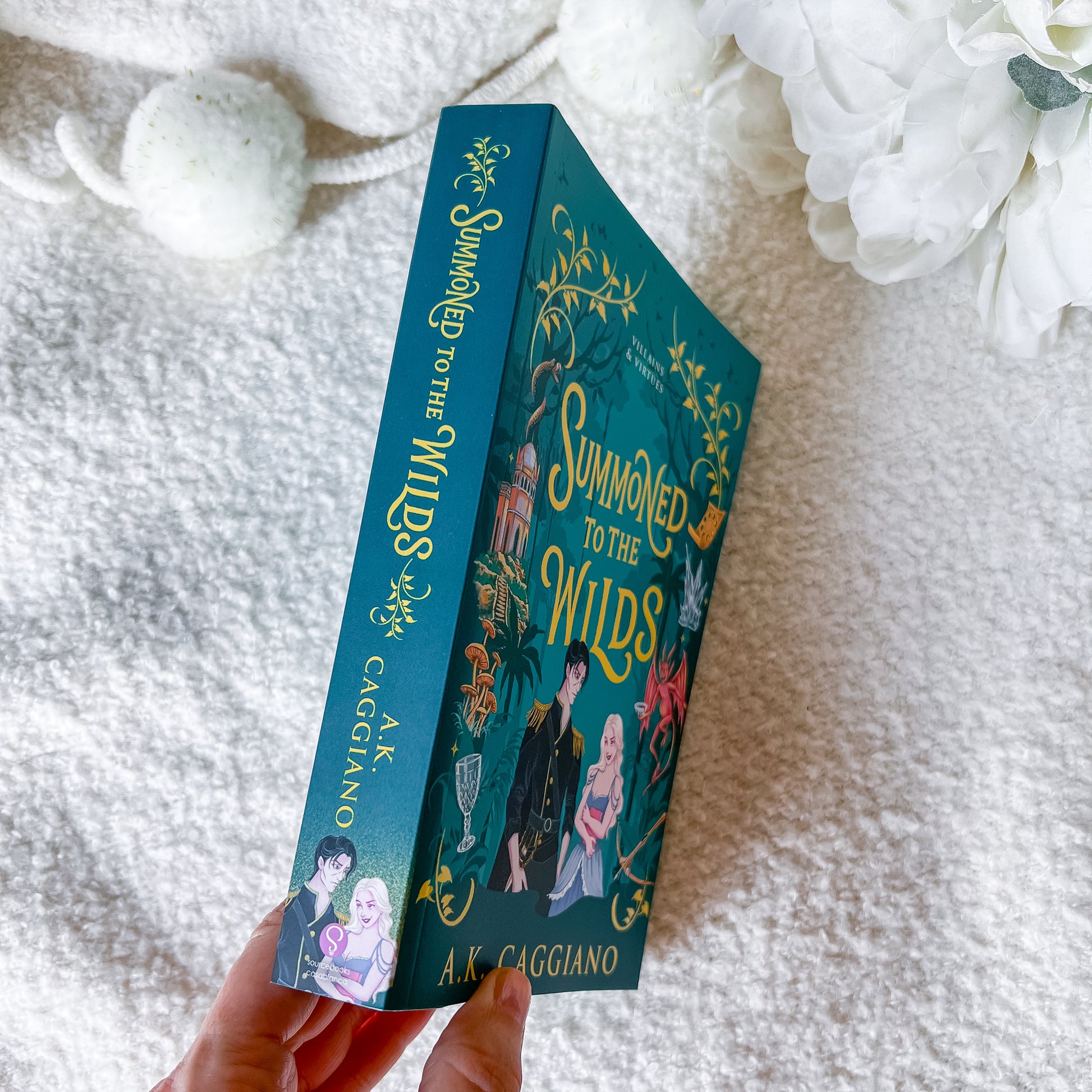 A hand holds a teal illustrated book titled "Summoned to the Wilds" by A.K.