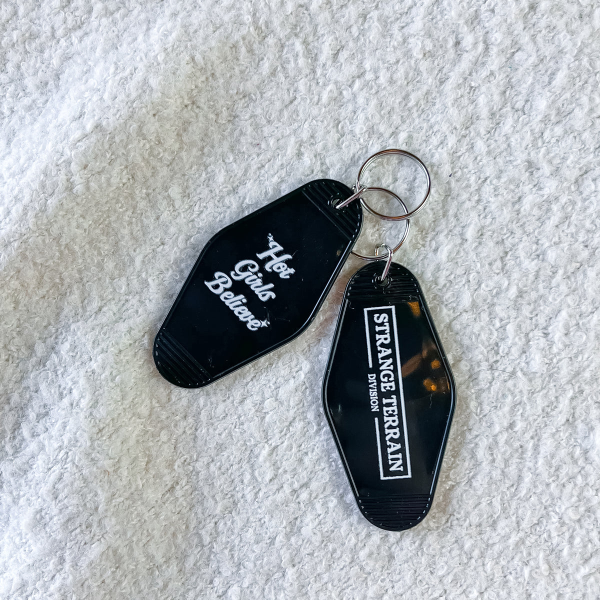 Two black acrylic keychains with silver keyrings on a textured white fabric background. One keychain is black with white script text reading 'Hot Girls Believe', and the other is black with a white border and text reading 'STRANGR TERRAIN DIVISION'.