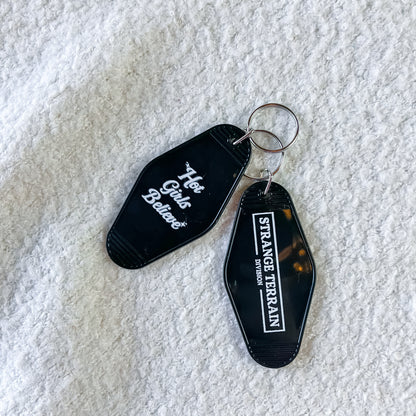 Two black acrylic keychains with silver keyrings on a textured white fabric background. One keychain is black with white script text reading 'Hot Girls Believe', and the other is black with a white border and text reading 'STRANGR TERRAIN DIVISION'.