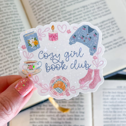 Sticker with 'Cozy girl book club' design held over an open book
