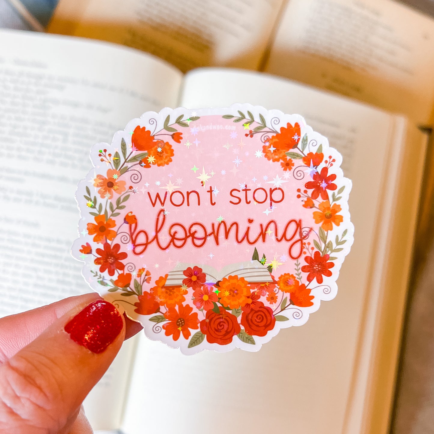 Sticker with floral design and 'won't stop blooming' text held over an open book.