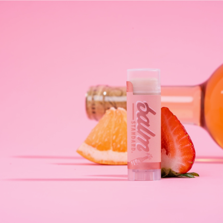 A pink lip balm tube labeled 'balmy' stands upright on a pink surface, styled with a slice of orange and a strawberry, with a pink bottle blurred in the background.