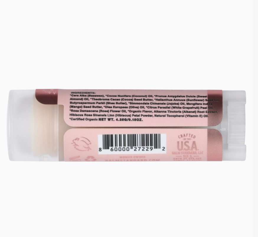 A single lip balm stick shown from the back, with the label facing up on a plain white background. The label is pink and white with a barcode and text including 'Crafted USA' and ingredient details.