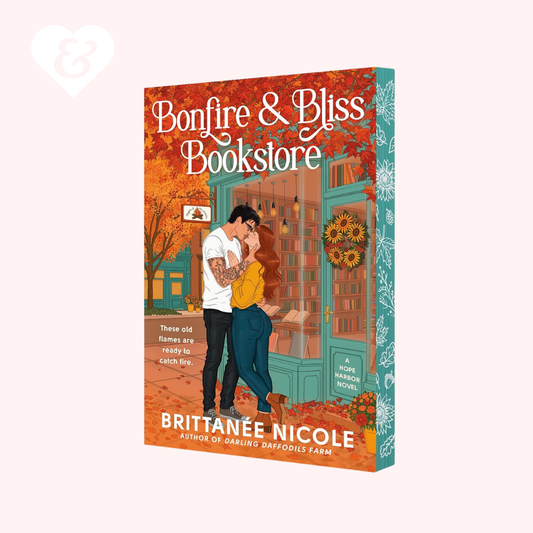 The book cover of 'Bonfire & Bliss Bookstore' by Brittanée Nicole, featuring an illustration of a couple in front of a bookshop with autumn foliage and teal storefront details, shown on a light pink background.
