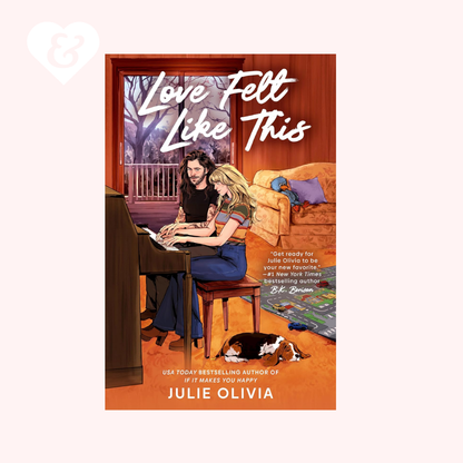 Book cover for 'Love Felt Like This' by Julie Olivia, featuring an illustrated scene of two women sitting at a piano in a warmly lit room, with a dog on the floor and a window showing an outdoor railing. The cover is centered on a pale pink background with a small white heart-and-ampersand mark in the upper left.