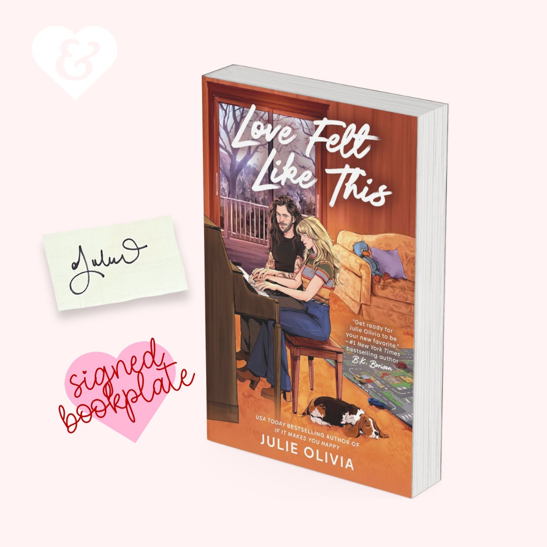A paperback copy of the book 'Love Felt Like This' by Julie Olivia is shown standing upright on a pale pink background, alongside a small white heart, a handwritten name card reading 'Julie', and a pink sign reading 'signed bookplate'. The cover features an illustrated couple in a warmly lit room with orange-brown tones and white title text.