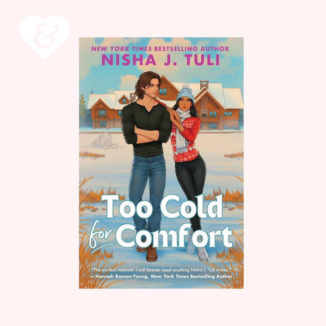 Book cover for 'Too Cold for Comfort' by Nisha J. Tuli, featuring an illustrated couple standing in a snowy outdoor setting with cabins in the background, shown on a pale pink background with a small white heart icon in the top-left corner.