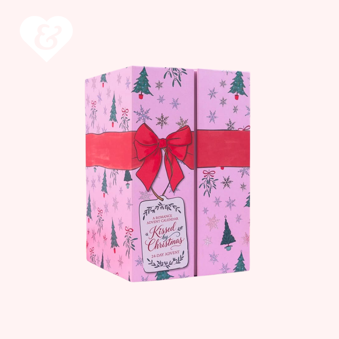 A pink Christmas-themed gift box with a winter tree and snowflake pattern, wrapped in a red ribbon with a large red bow and a hanging gift tag that reads 'Kissed & Christmas'.