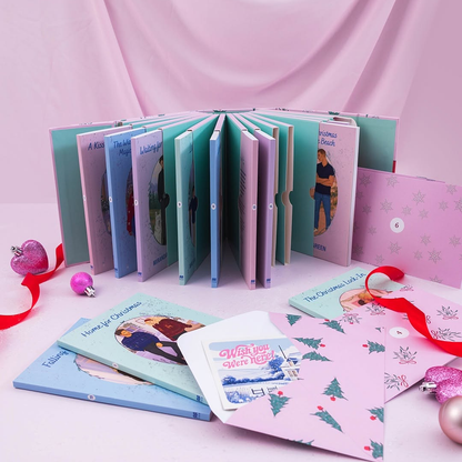 A pastel Christmas-themed book or album set arranged on a pink surface with a pink draped backdrop. The set includes multiple soft-cover books and envelopes/cards with illustrated figures, Christmas trees, snowflakes, and red ribbon accents.