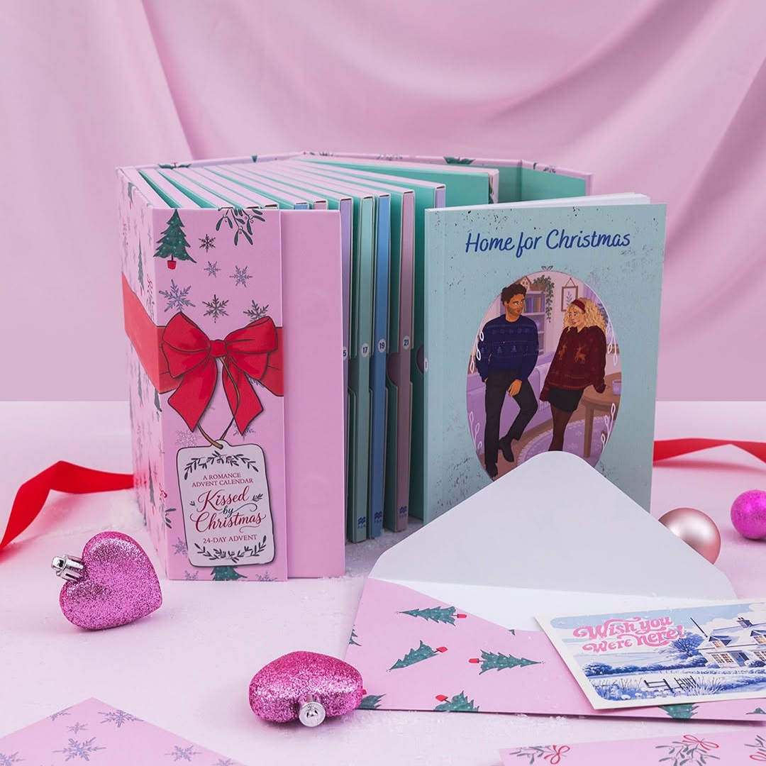A boxed set of Christmas-themed greeting cards or booklets arranged on a pink surface with a pink draped backdrop. The set includes pastel pink and mint volumes, a red ribbon bow on the front, an open white envelope, a pink glitter heart-shaped ornament, and a holiday card reading 'Home for Christmas.