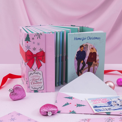A boxed set of Christmas-themed greeting cards or booklets arranged on a pink surface with a pink draped backdrop. The set includes pastel pink and mint volumes, a red ribbon bow on the front, an open white envelope, a pink glitter heart-shaped ornament, and a holiday card reading 'Home for Christmas.