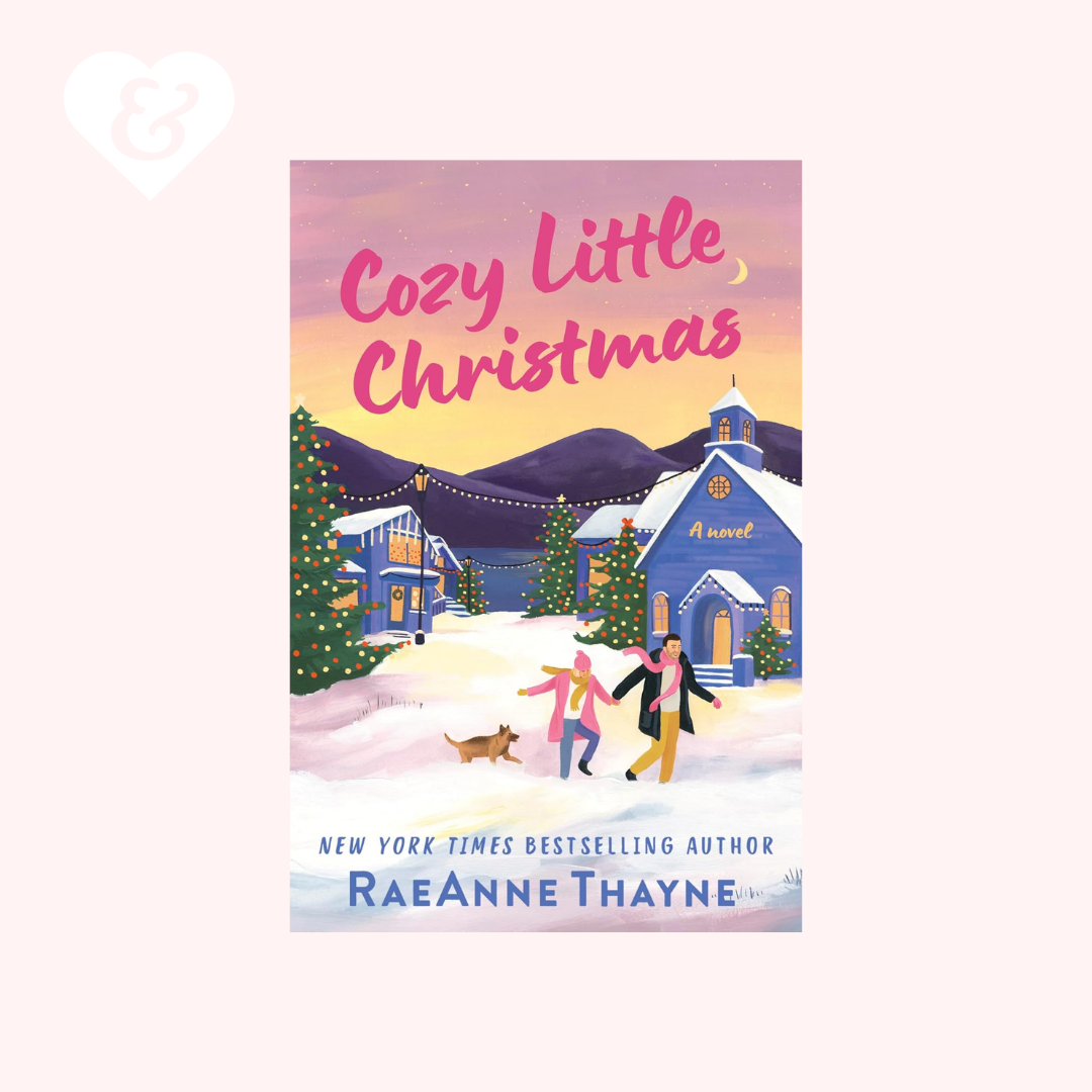 Book cover for 'Cozy Little Christmas' by RaeAnne Thayne, featuring a winter village scene with snow-covered houses, decorated trees, and two people walking in the snow. The cover is centered on a pale pink background with a small white heart-and-ampersand mark in the top-left corner.