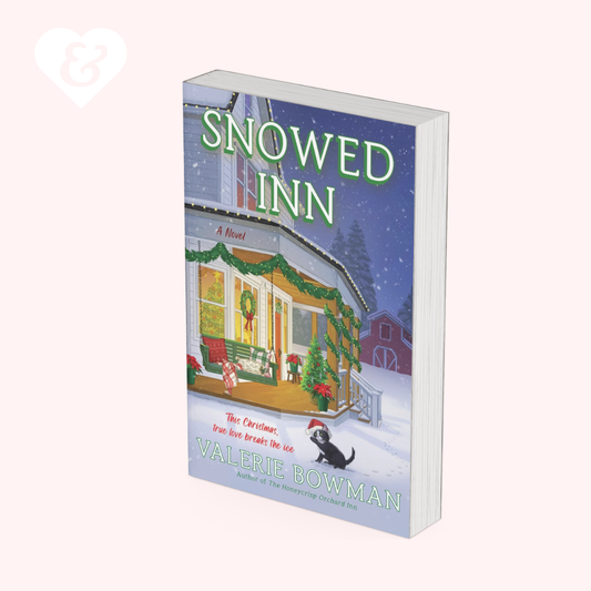 The book cover of 'Snowed Inn' by Valerie Bowman, shown as a 3D mockup on a pale pink background. The cover features a festive Christmas night scene of a snow-covered inn with green garlands, a black cat in a Santa hat, and a snowy landscape under a dark blue sky.