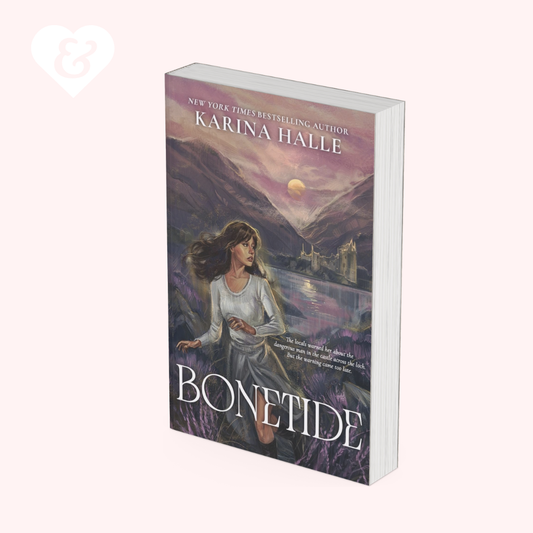 A paperback copy of the book 'Bonetide' by Karina Halle, featuring a fantasy cover illustration of a woman in a white dress standing in a field of purple flowers near a lake and castle under a sunset sky. The book is shown standing upright against a pale pink background.