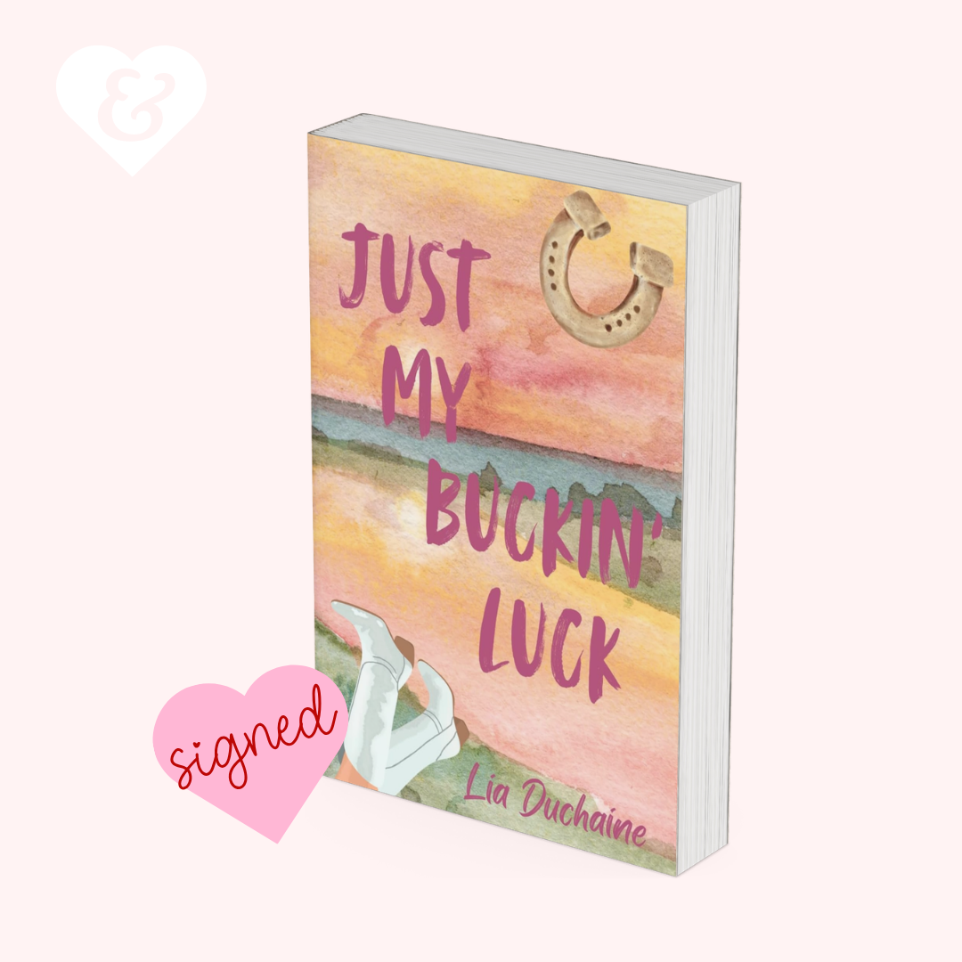 A 3D mockup of a book titled 'Just My Buckin' Luck' by Lia Duchaine, featuring a pastel watercolor cover with pink lettering, a horseshoe, and white high heels. The book is displayed on a light pink background with a white heart icon and a pink 'signed' heart sticker.