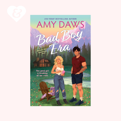 Book cover for 'Bad Boy Era' by Amy Daws, featuring an illustrated blonde woman in ripped jeans and a man in a maroon shirt standing in a mountain landscape with a cabin and lake. The cover is centered on a pale pink background with a faint heart icon in the top left.