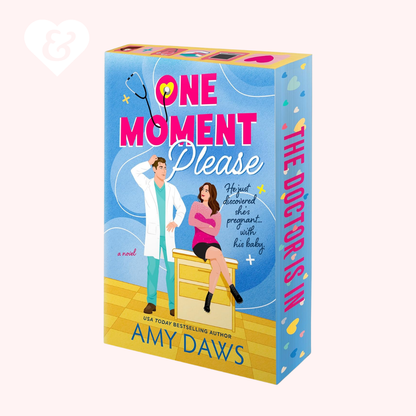 A boxed book cover for the novel 'One Moment Please' by Amy Daws, shown at a slight angle on a pale pink background. The box features a bright blue front with pink title text, an illustration of a doctor and patient, and a yellow floor, plus the spine text 'THE DOCTOR IS IN'.