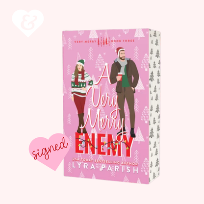 A boxed book cover for 'A Very Merry Enemy' by Lyra Parish, shown on a pale pink background with decorative heart graphics. The box is predominantly pink with illustrated holiday characters and white tree patterns.