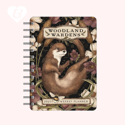 A spiral-bound 2027 weekly planner titled 'Woodland Wardens' featuring an illustration of an otter among flowers and leaves on a pale pink background.