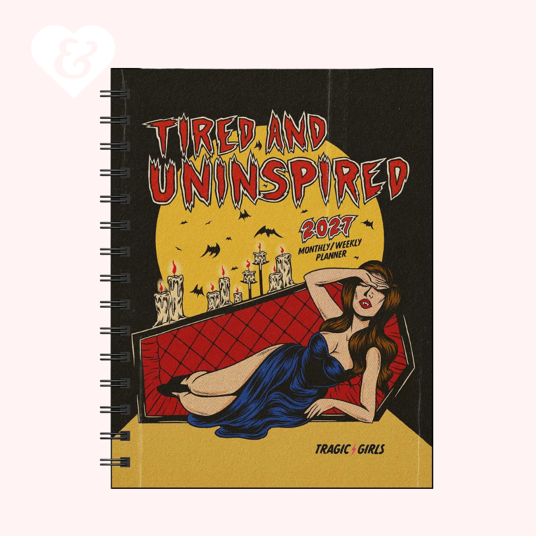 A spiral-bound weekly planner titled 'Tired and Uninspired 2023 Monthly Weekly Planner' with a gothic pin-up illustration of a woman in a blue dress reclining in a red coffin against a yellow moon, shown on a pale pink background.