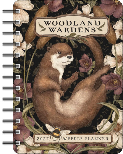 Woodland Wardens 2027 Weekly Planner notebook featuring an illustrated brown otter surrounded by flowers and leaves on a dark background, with spiral binding visible along the left side.